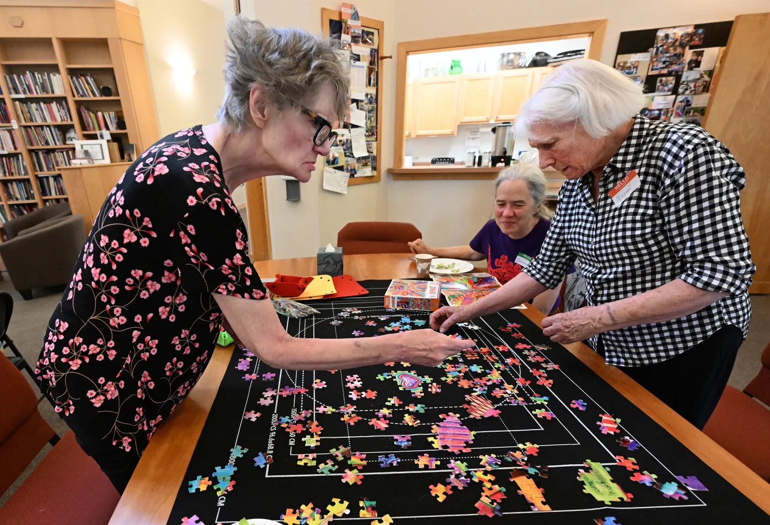 Three women sit and stand around a table working on a colorful jigsaw puzzle together at Recovery Café Longmont's Boulder Pop-Up