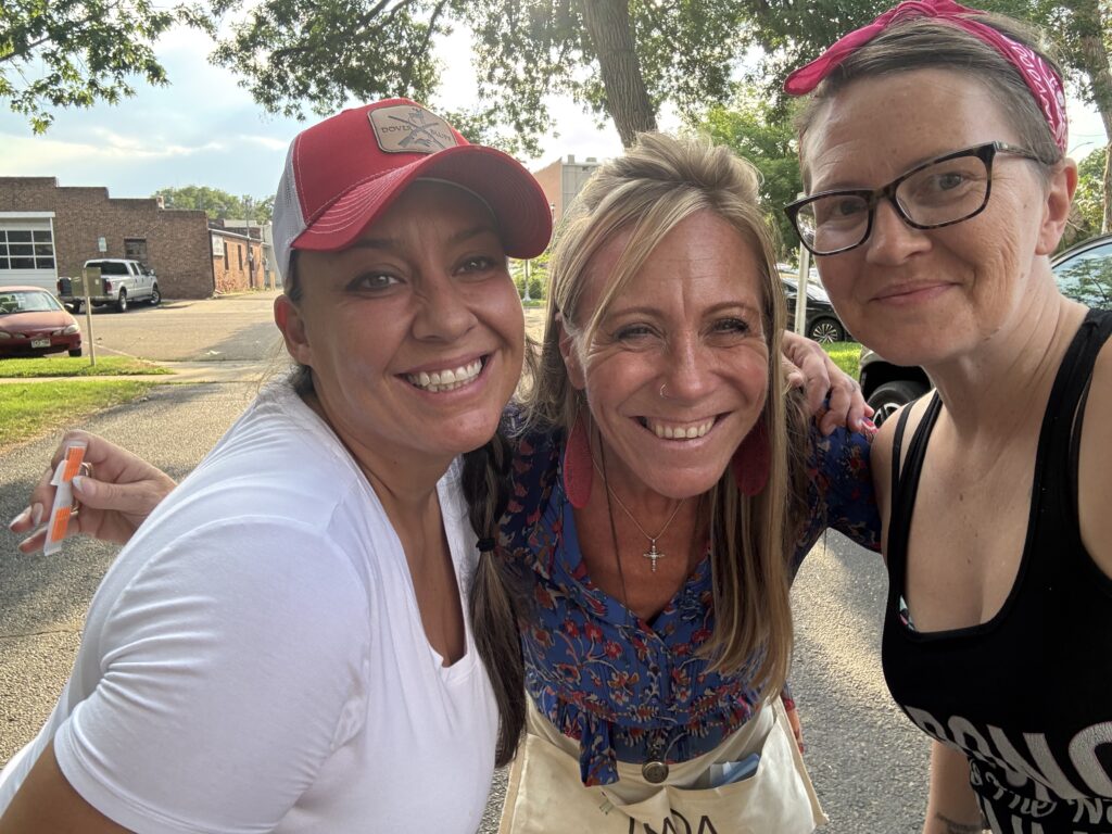 Three smiling women stand close together outdoors, enjoying a sunny day at a Recovery Café Longmont community event.