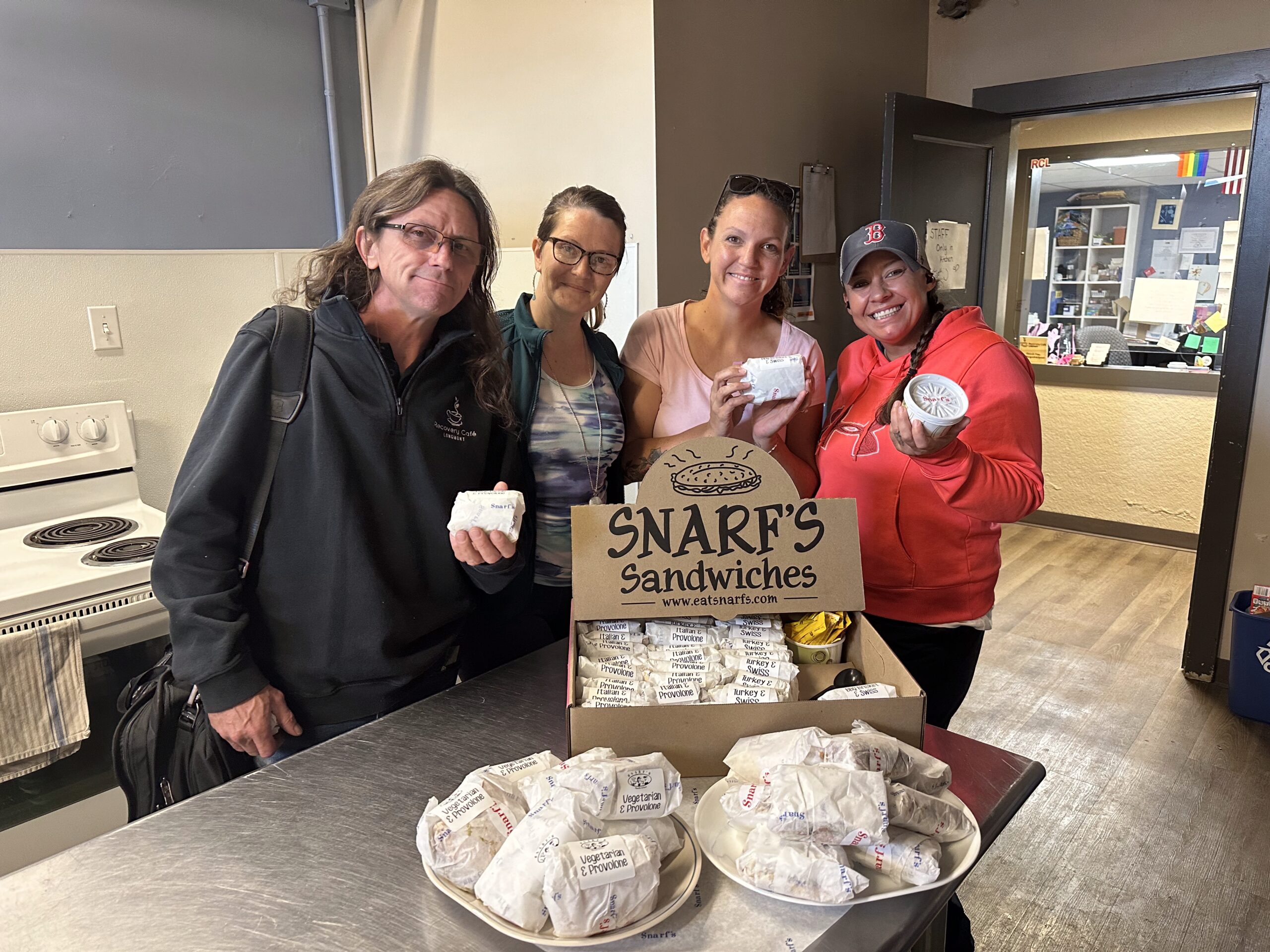Four people at Recovery Café Longmont smile while holding donated Snarf’s sandwiches in the Café kitchen, highlighting community and generosity.