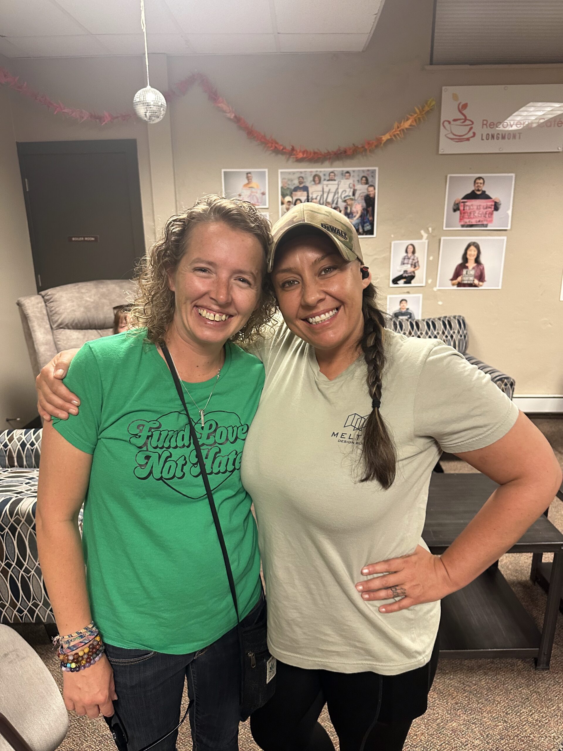 Two women smile and embrace inside Recovery Café Longmont, standing in front of a wall of photos and community art.