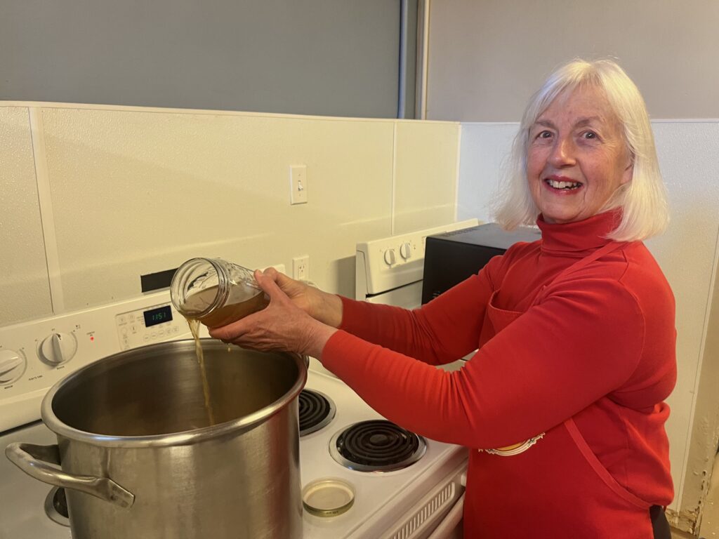 A recovery Café Longmont volunteer with white hair and a red turtleneck pours a liquid into a stock pot, making a delicious nutritious meal for the community.
