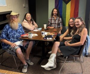 Five people sit around a wooden table sharing food and conversation at Recovery Café Longmont. A colorful pride flag hangs on the wall behind them.