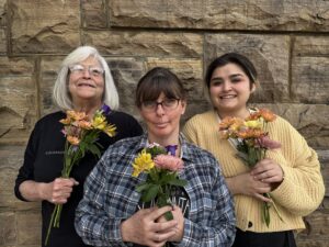 Group of three recovery Café Longmont members welcome new guests, and visitors with flowers. They represent new beginnings and joy in the Café. 