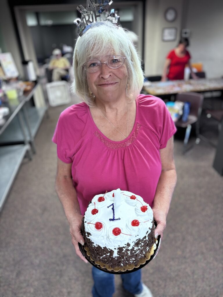 A smiling woman with white hair and glasses holds a chocolate cake topped with white frosting, cherries, and a large “1” candle. She’s wearing a bright pink shirt and a silver “Happy Birthday” tiara.